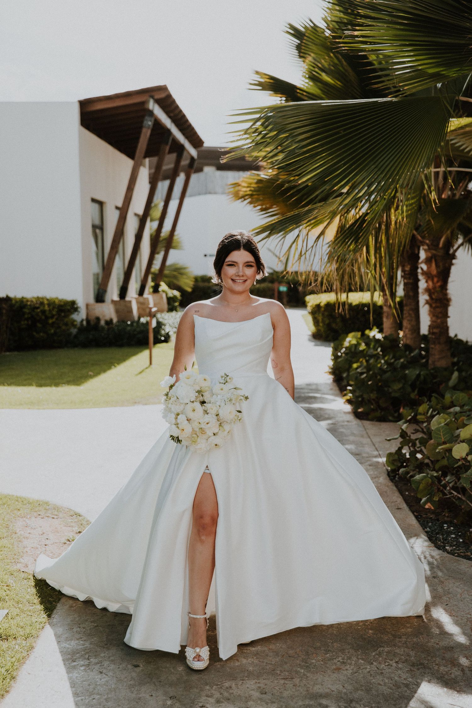 Dreams Macao beach  bride in a flowing white gown with thigh-high slit holds a white bouquet against tropical scenery.