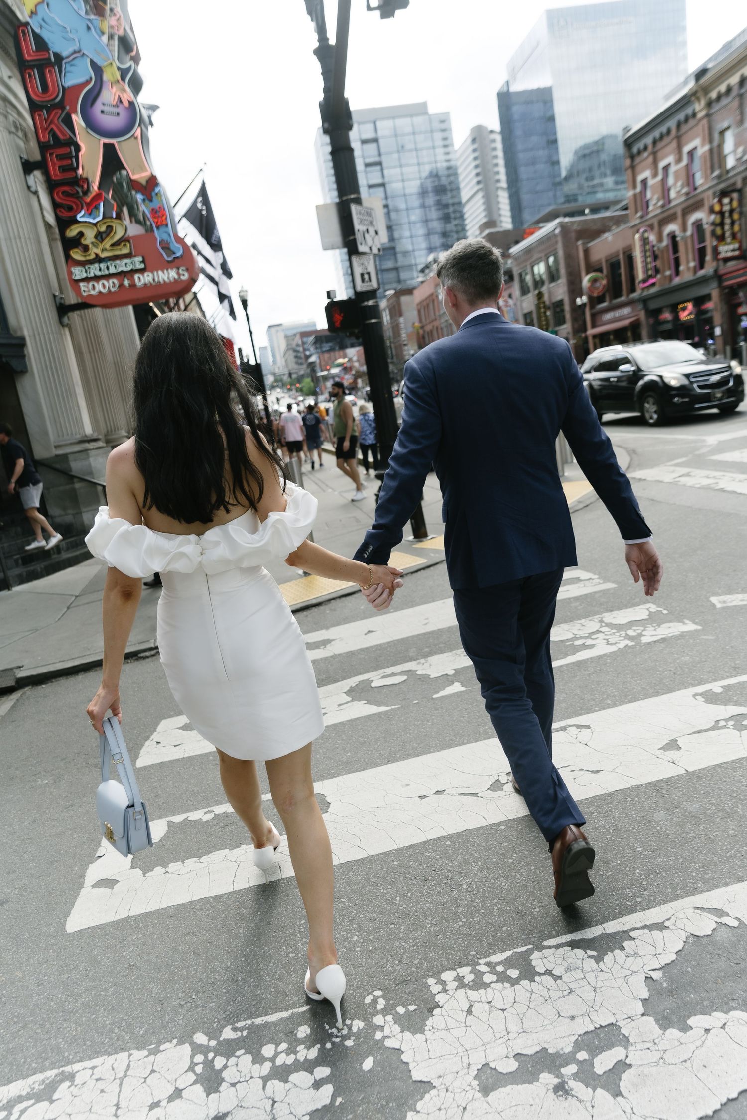 A couple walking across a crosswalk in downtown, with the woman in white dress and man in navy suit.