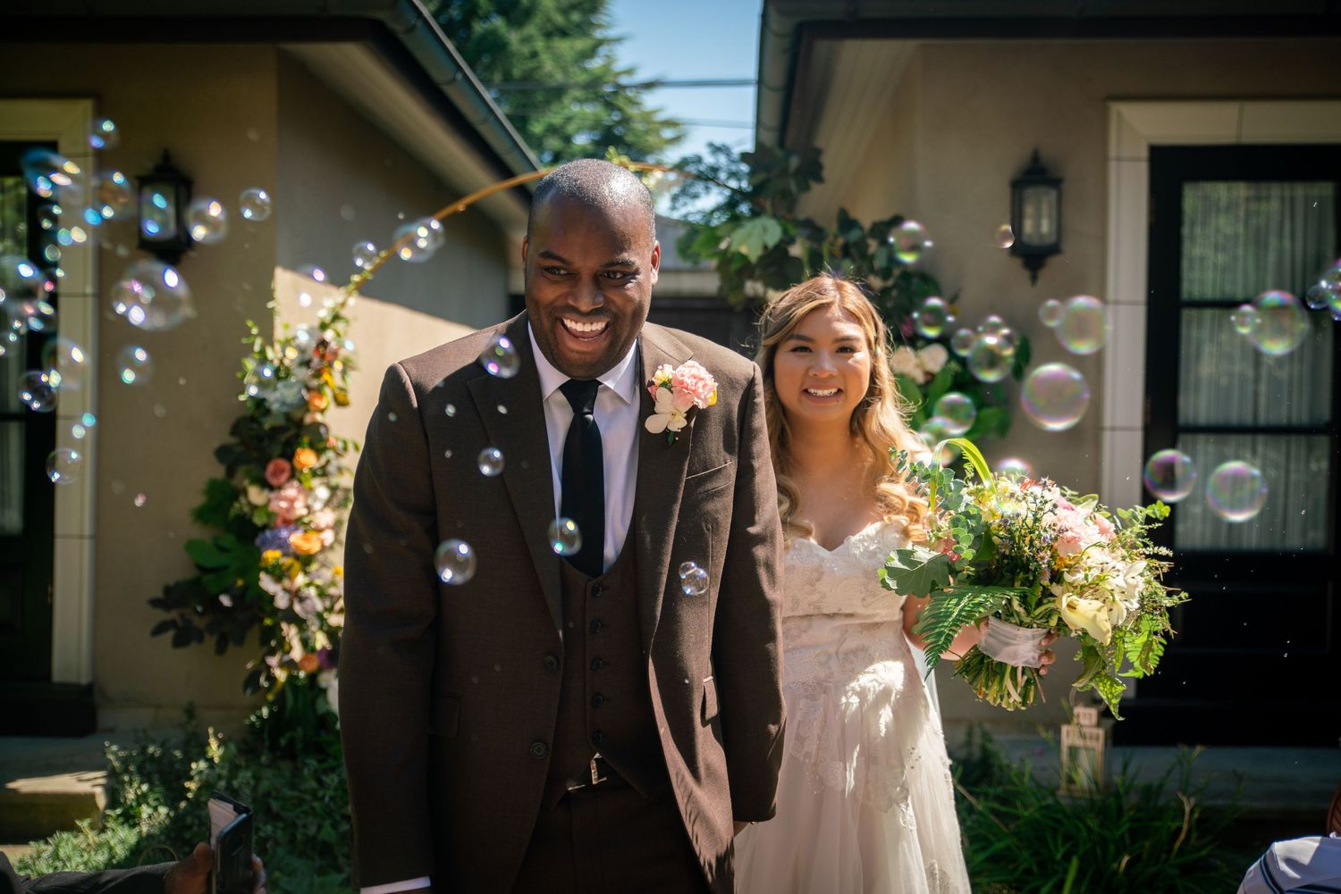 A couple celebrates their wedding day outdoors with bubbles floating in the sunlit air.