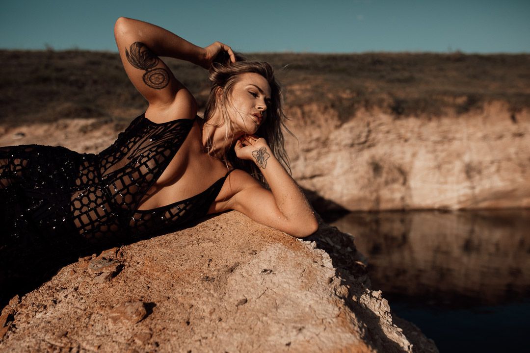 Model in black lace dress poses elegantly on rocky cliff edge beside calm water at sunset.