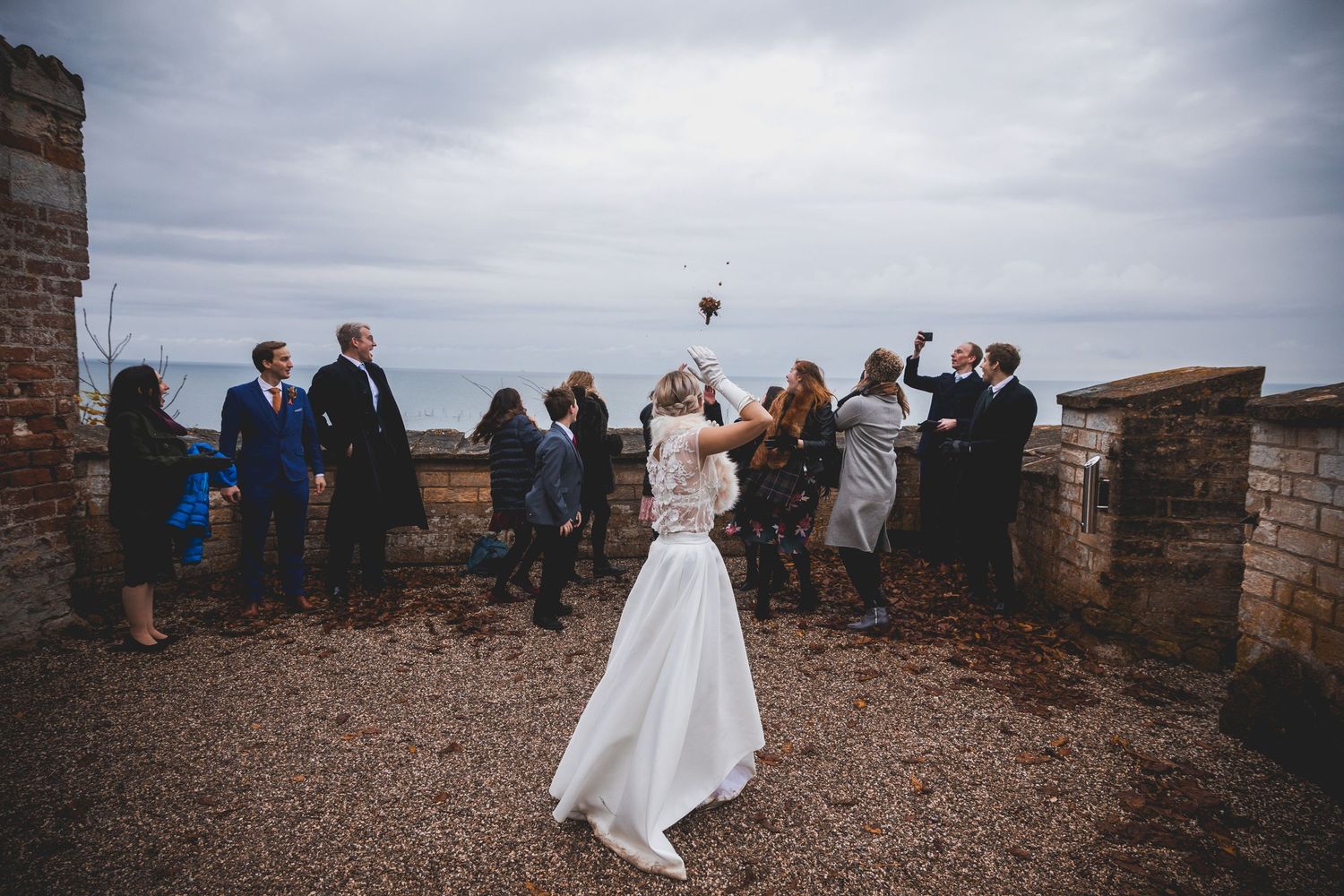 Scenic wedding photo on castle terrace with guests and dramatic cloudy sky.