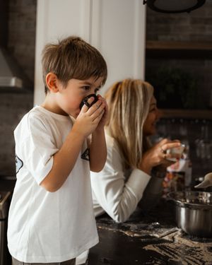 A child in white sips from a mug while baking in a cozy kitchen setting.