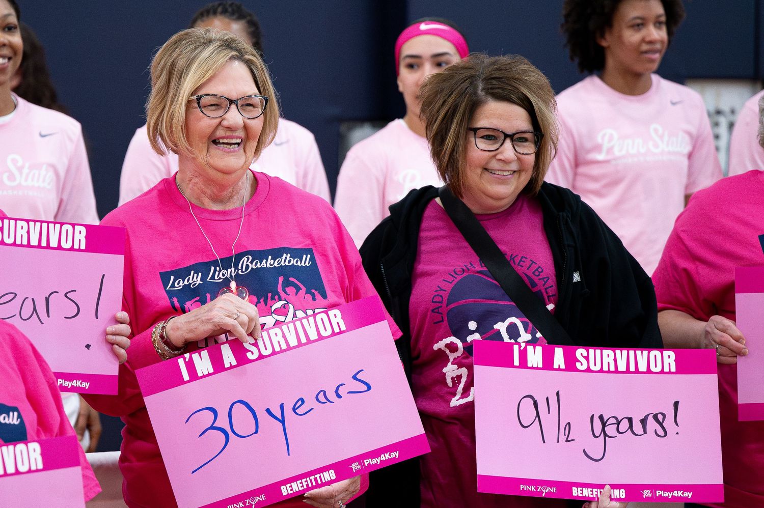 Cancer survivors in matching pink shirts celebrate together displaying signs showing their years of survival.