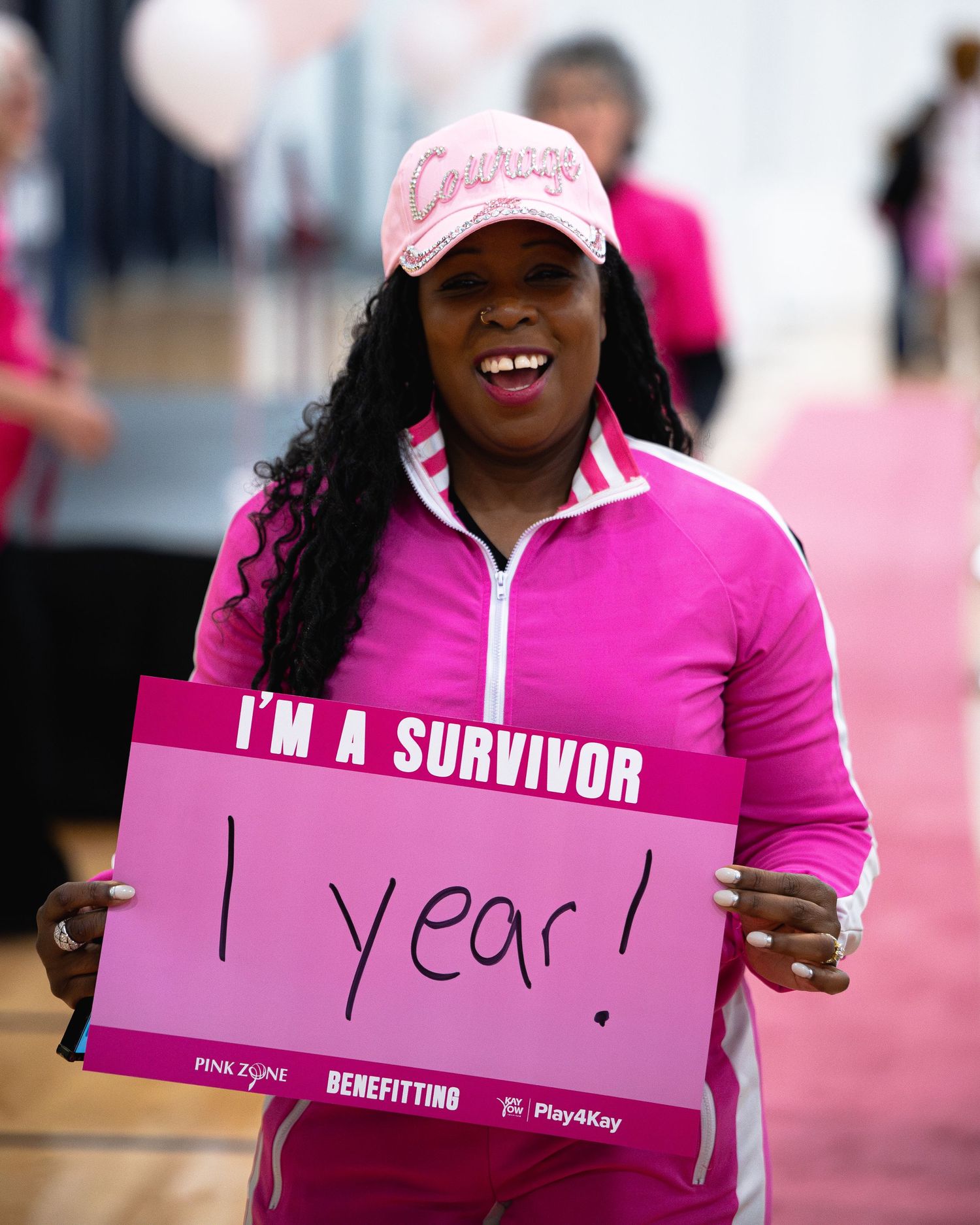 Cancer survivor in pink jacket joyfully holds sign celebrating one year of survival.