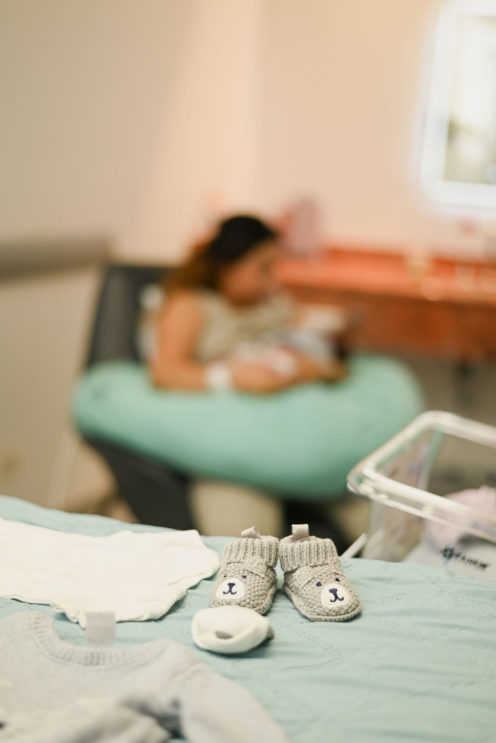Baby booties and clothing lay on a mint green surface while someone rests on a nursing pillow in the background.