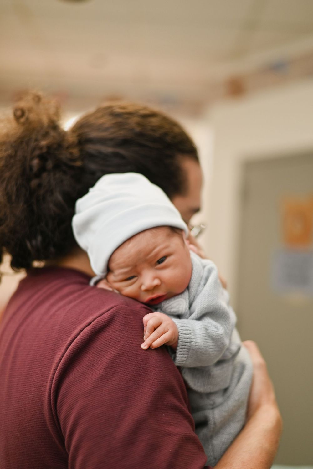 A sleeping newborn baby wearing a white hat and gray sweater rests peacefully on someone's shoulder.