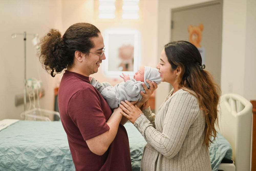 Two people in a bedroom share a sweet moment as they hold a baby together wearing burgundy and beige sweaters.