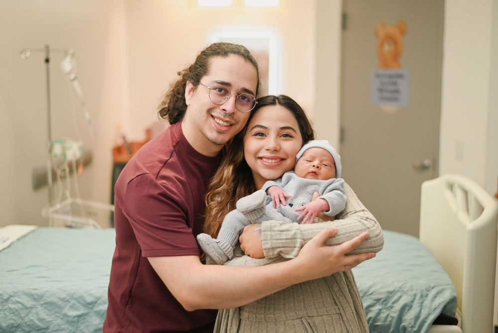 A happy couple holds their newborn baby while sitting together in a hospital room with medical equipment nearby.