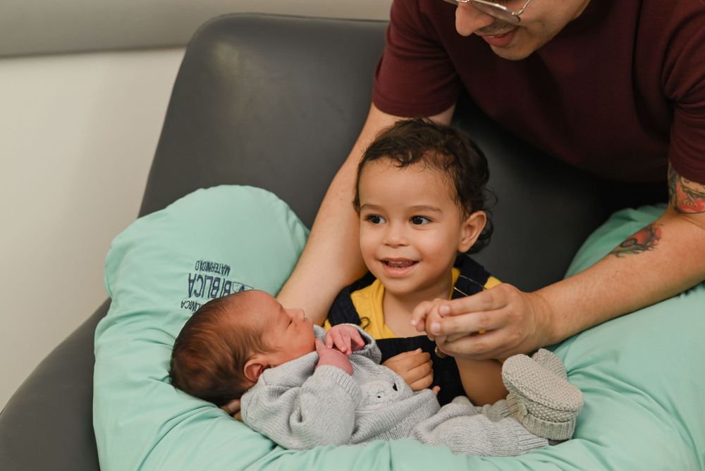 Two young children sit together on a mint green nursing pillow while an adult helps them interact.