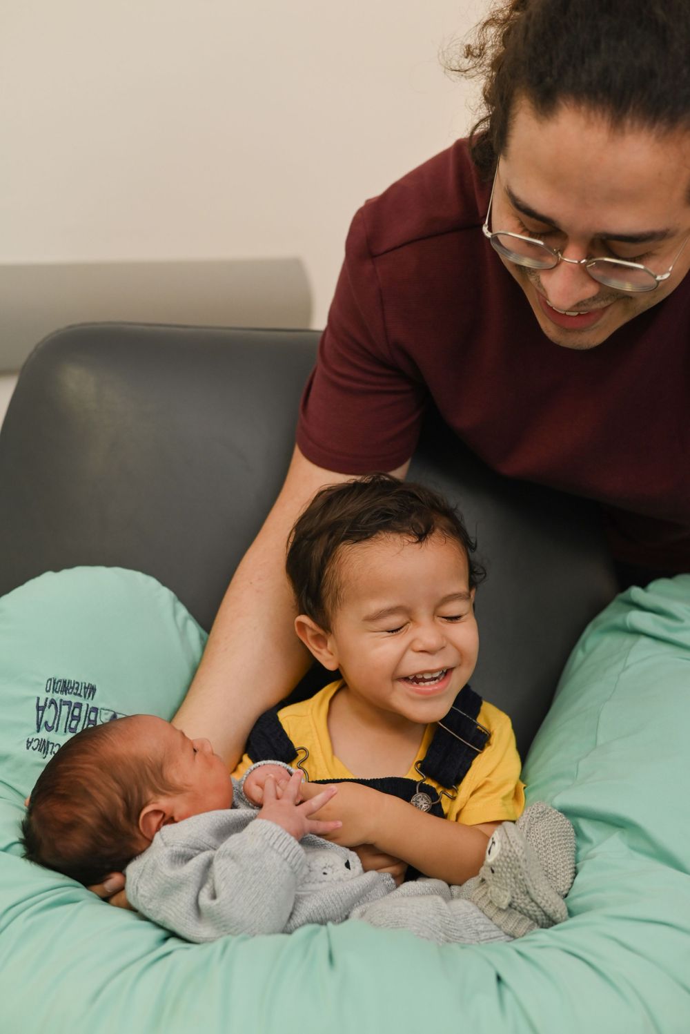 A child in a yellow shirt laughs joyfully while looking at a newborn baby resting on mint green bedding.