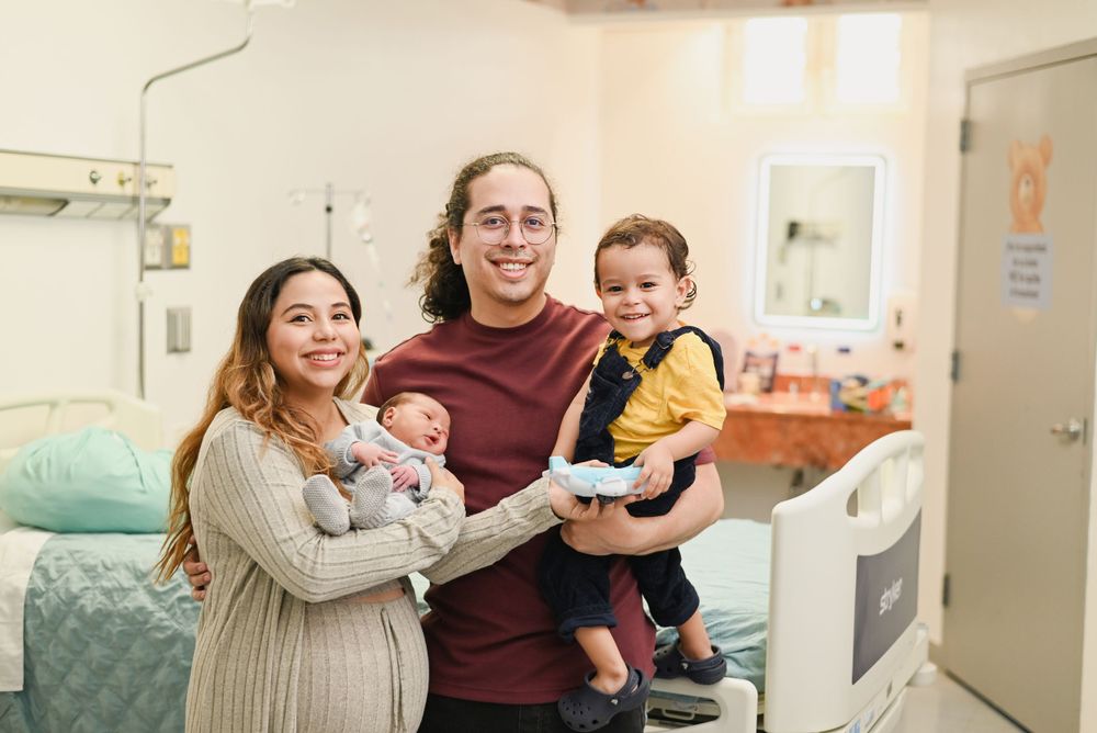 A smiling family poses together in a medical room with a baby and toddler, showing warmth and joy.