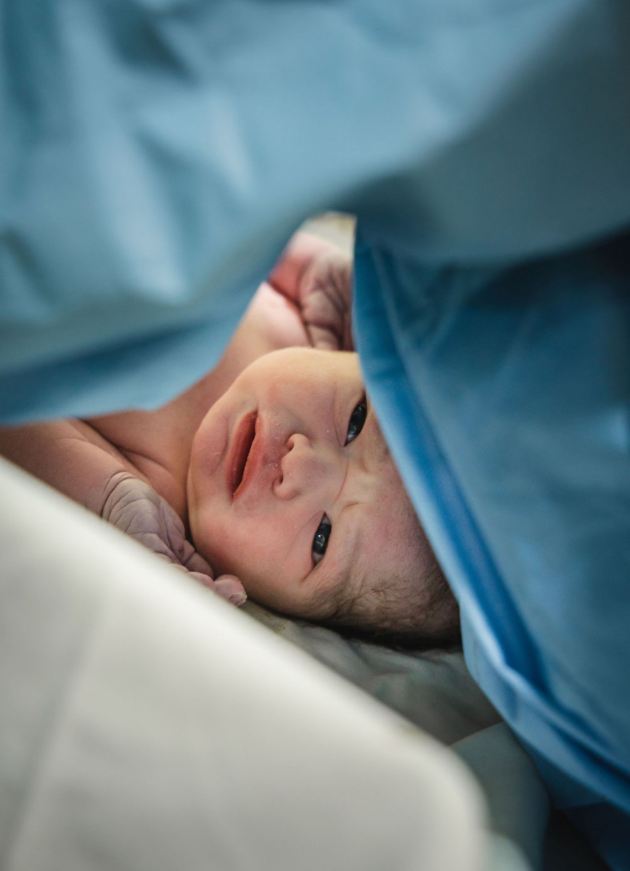 Baby lying in hospital bed smiles while surrounded by blue medical curtains and white sheets.
