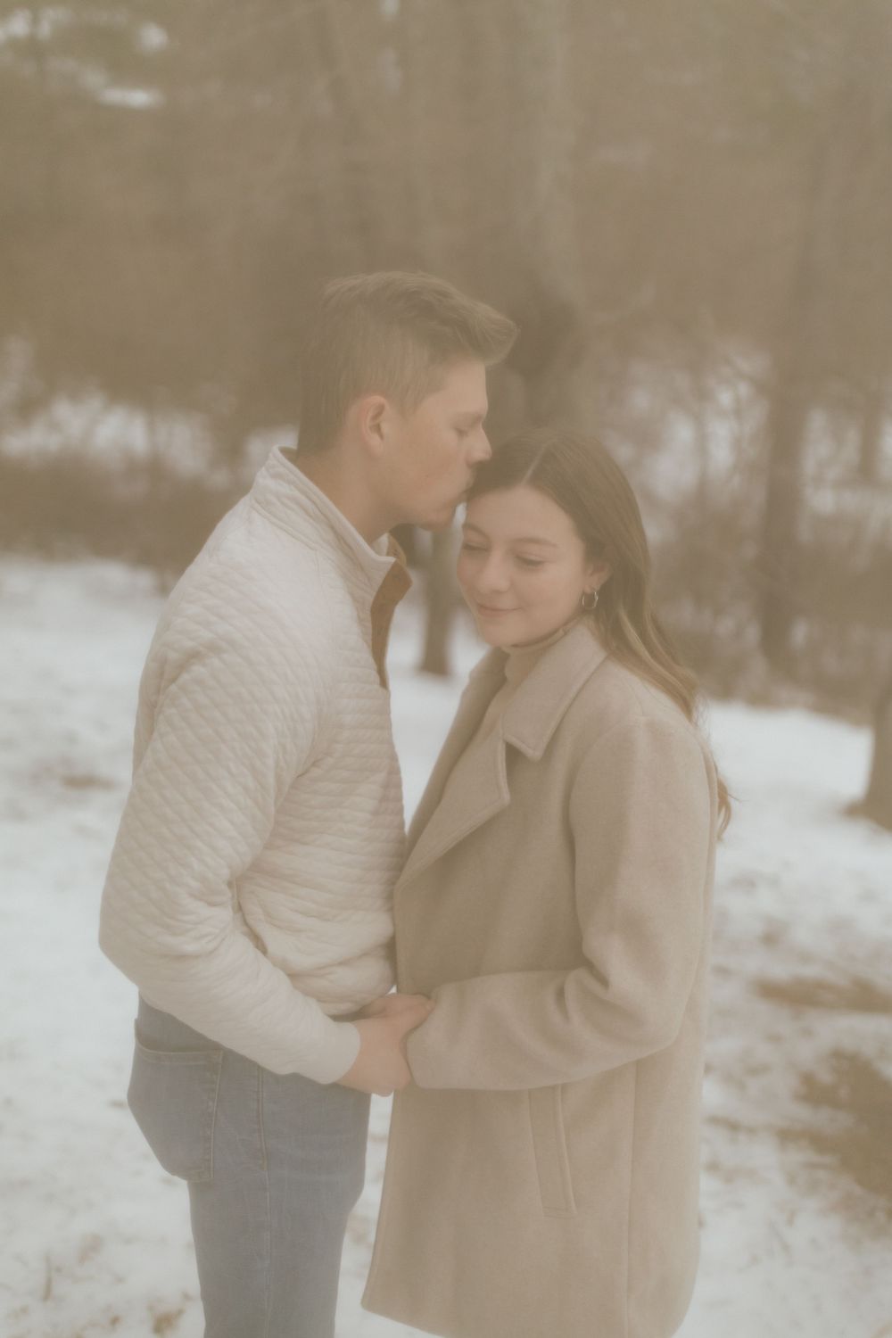 Intimate moment between couple in neutral winter clothing against snowy forest backdrop.