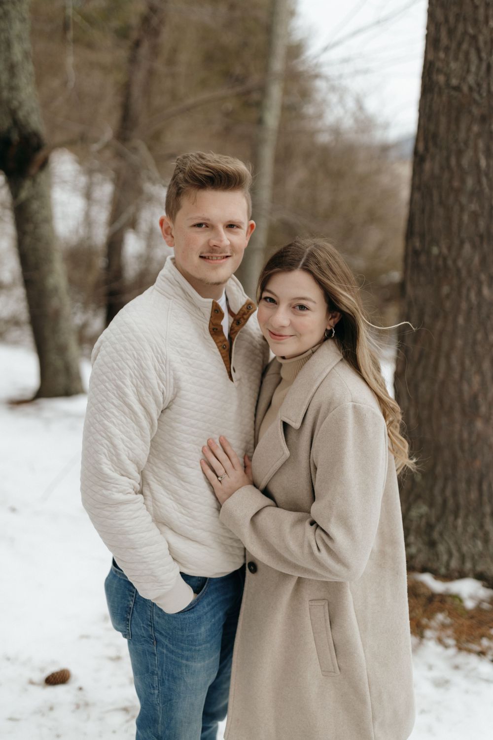 Winter engagement photo of couple in coordinating neutral outfits standing among trees.