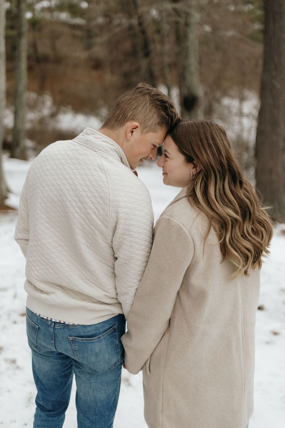 Intimate winter portrait of couple in cream sweaters sharing tender moment in snowy woods.