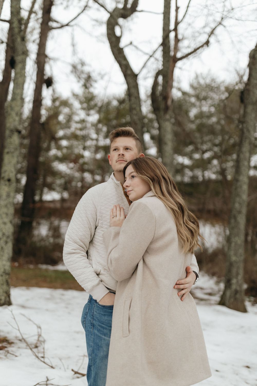 Romantic winter engagement photo of a couple hugging in a snowy forest setting.