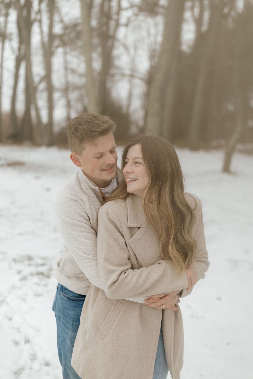 A romantic winter engagement photo shows a couple embracing in a snowy forest setting while wearing coordinating beige outfits.
