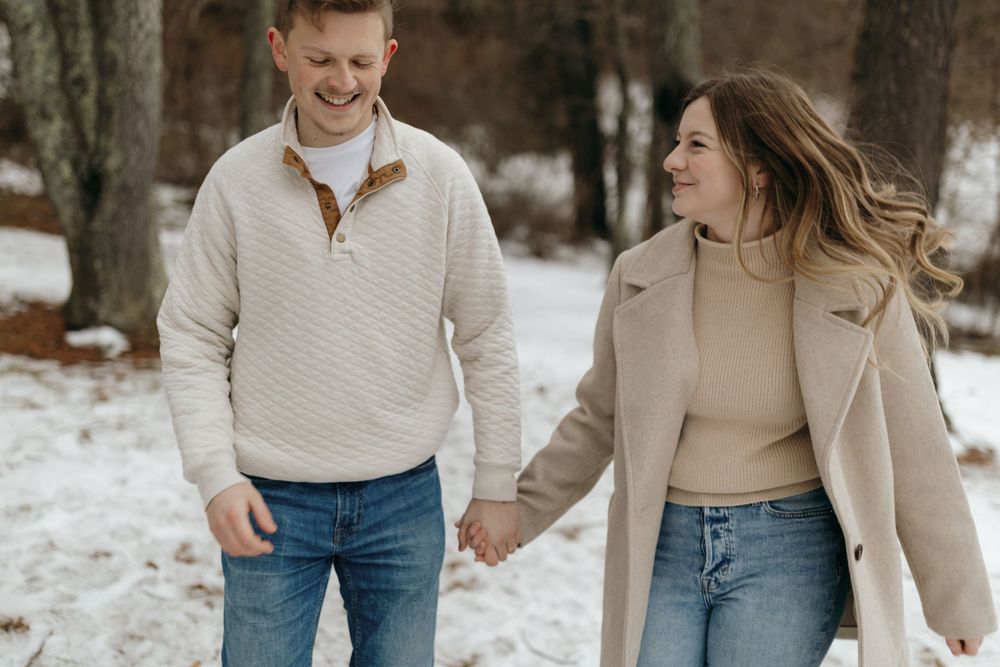 A couple walking together in the snow wearing matching neutral tones and sharing a joyful moment.