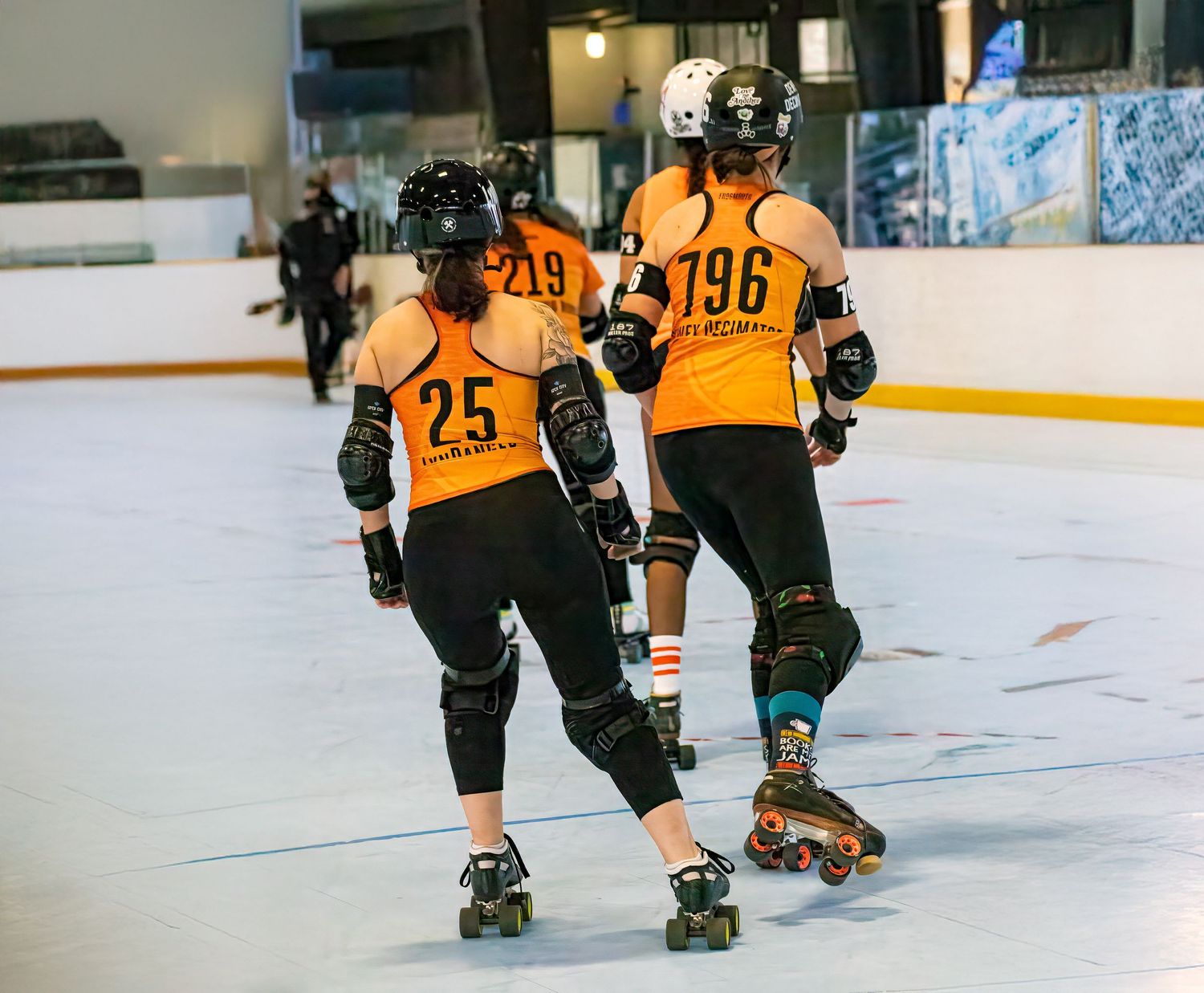Four roller derby players in orange jerseys skate on an indoor rink.