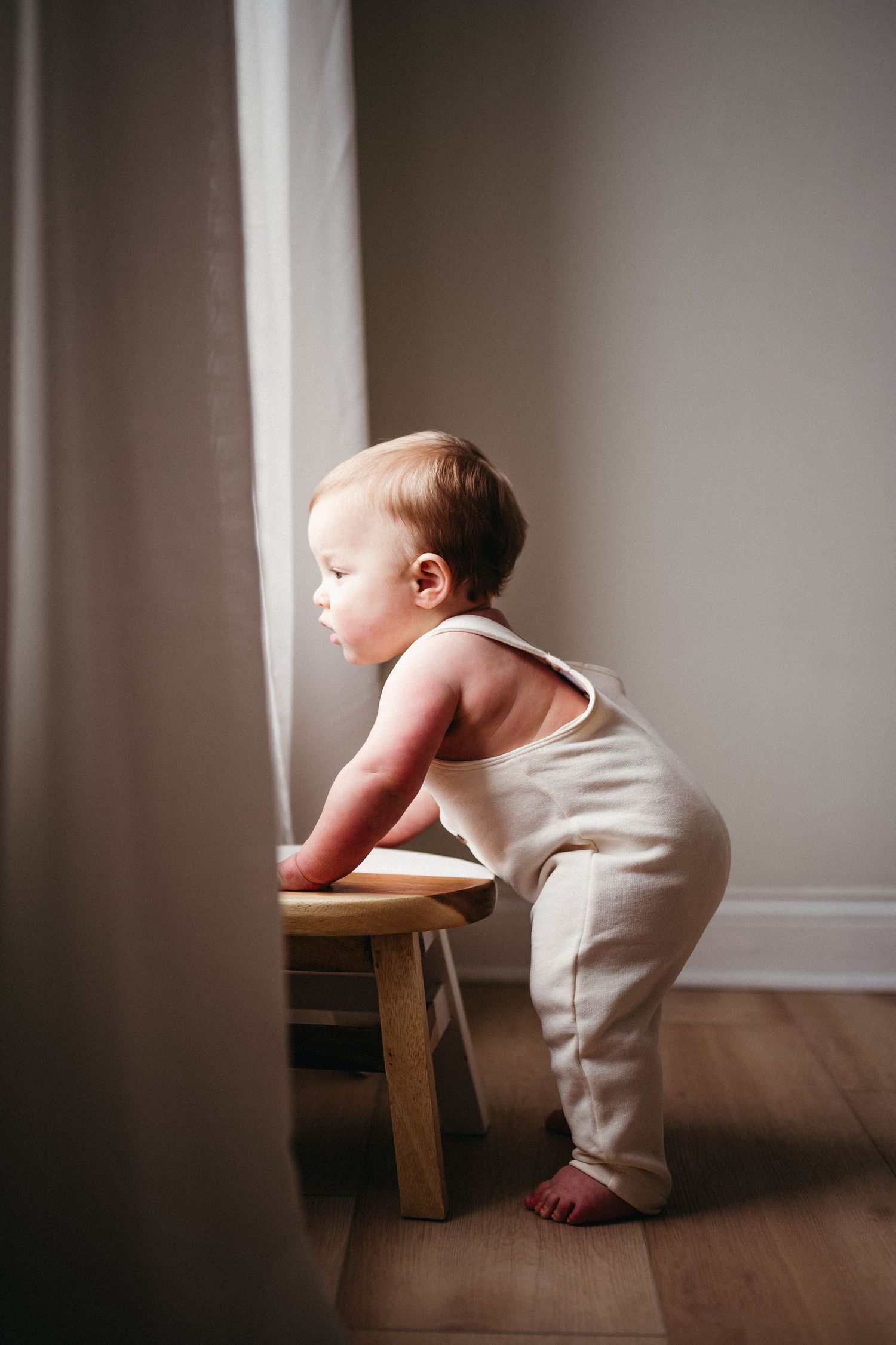 Baby in white romper reaching up to windowsill while learning to stand.