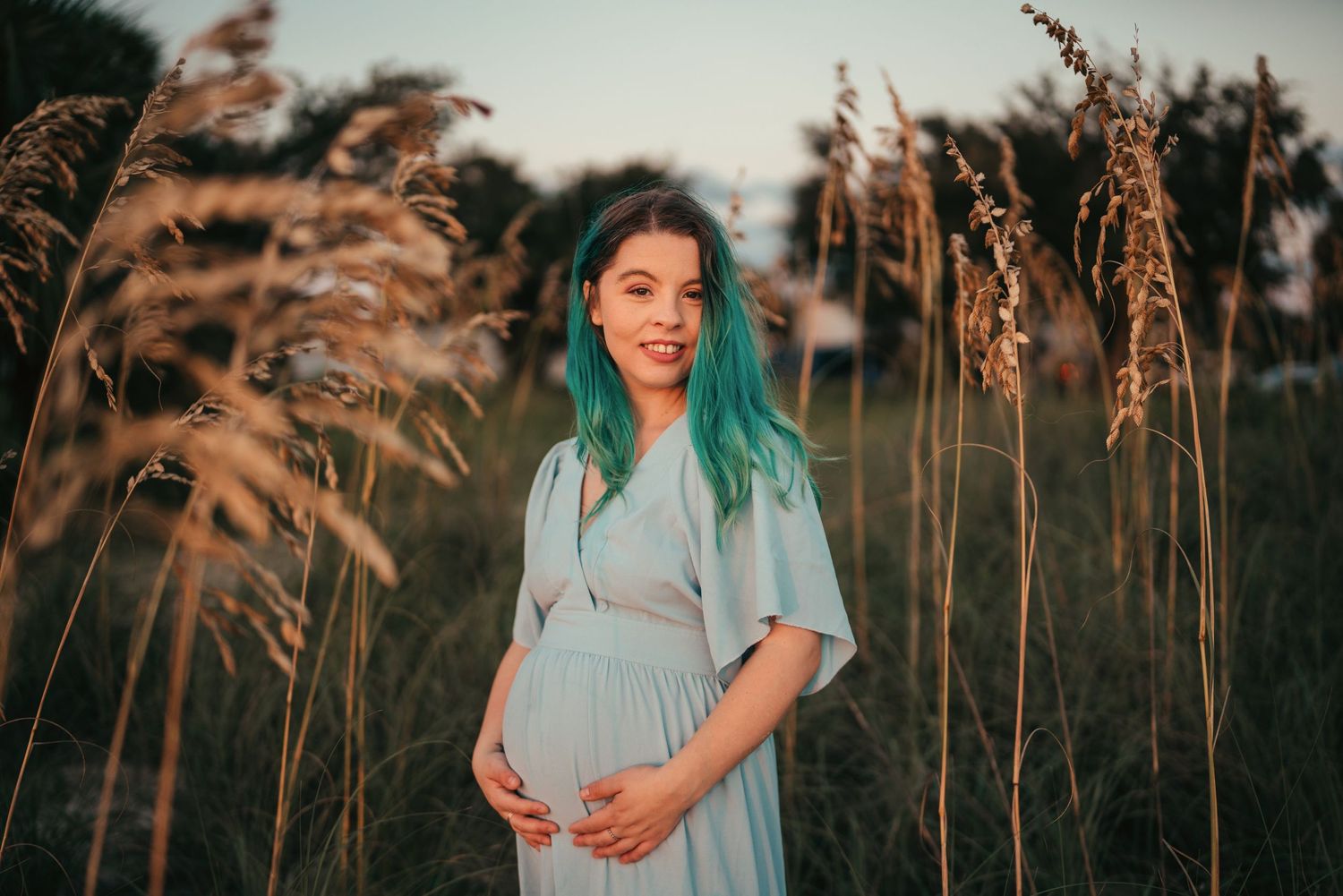 Expectant woman in blue dress stands among tall golden grasses at sunset with teal-colored hair.