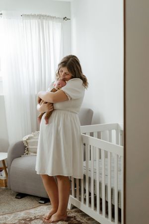 A parent in a white dress holding a sleeping baby in a minimalist nursery.