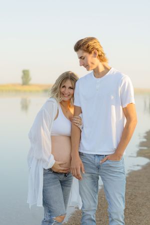 A couple stands together by a serene lake at sunset.