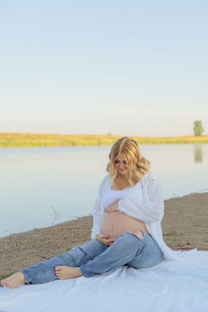 Maternity photo of woman sitting on white blanket by lake during sunset.