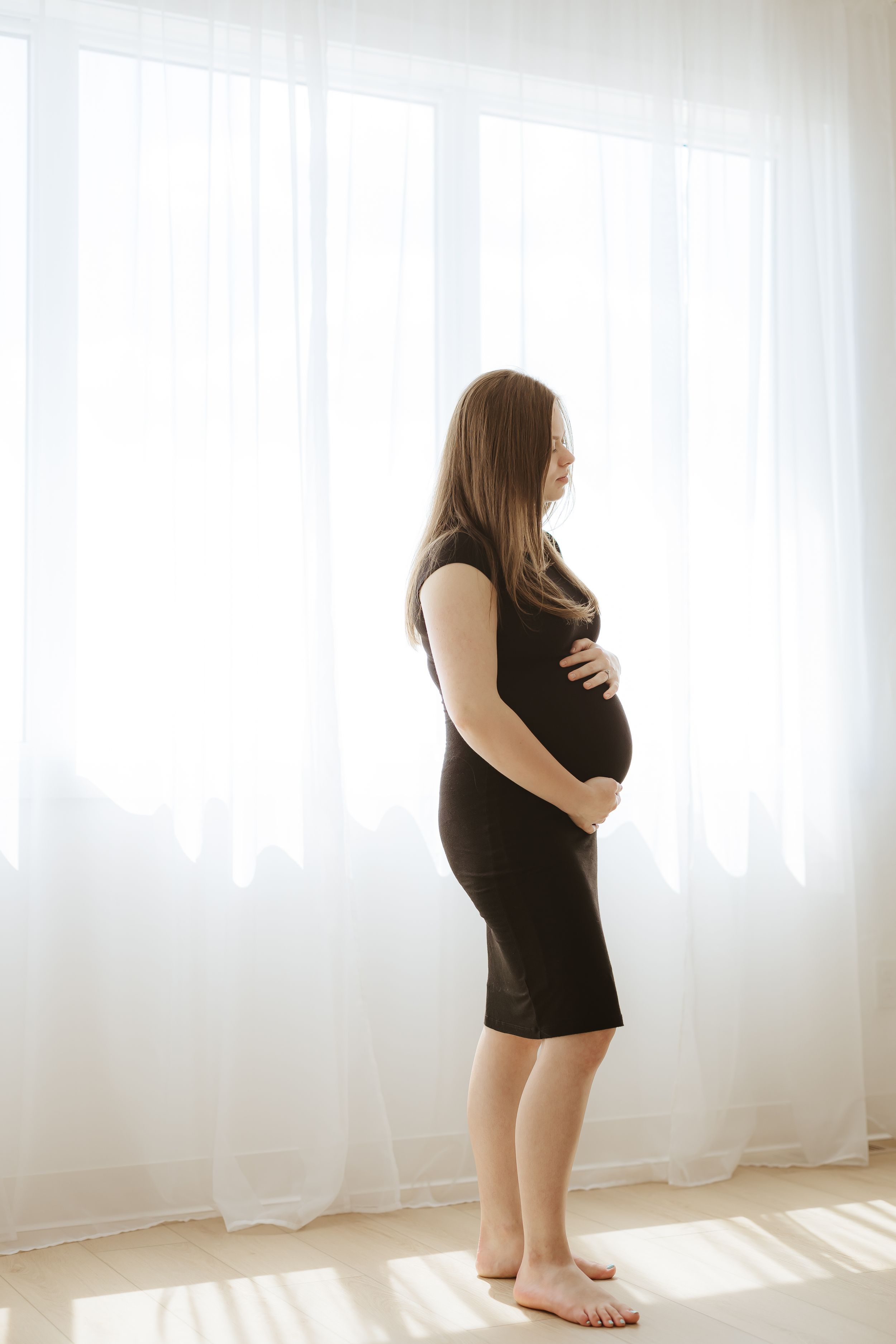 A silhouette of an expectant person in a black dress stands barefoot against a bright window.