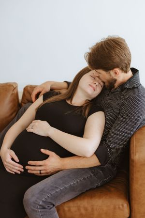 Intimate couple rests together on a brown leather couch, sharing a tender moment during pregnancy.