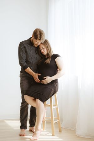 An intimate maternity moment captured with a couple by a bright window and wooden stool.