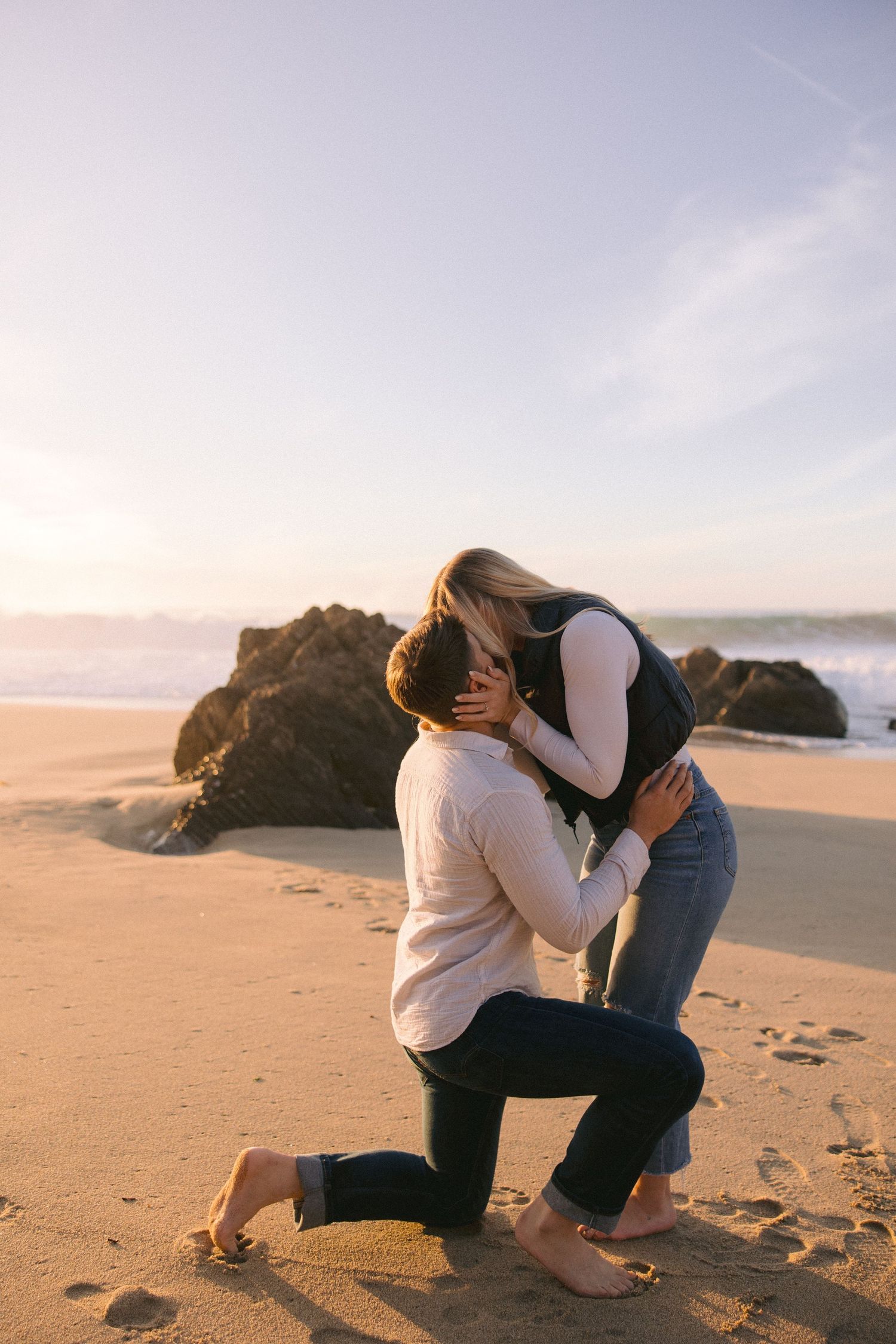 Two people share an intimate moment on a sandy beach at sunset with rocky outcroppings in the background.