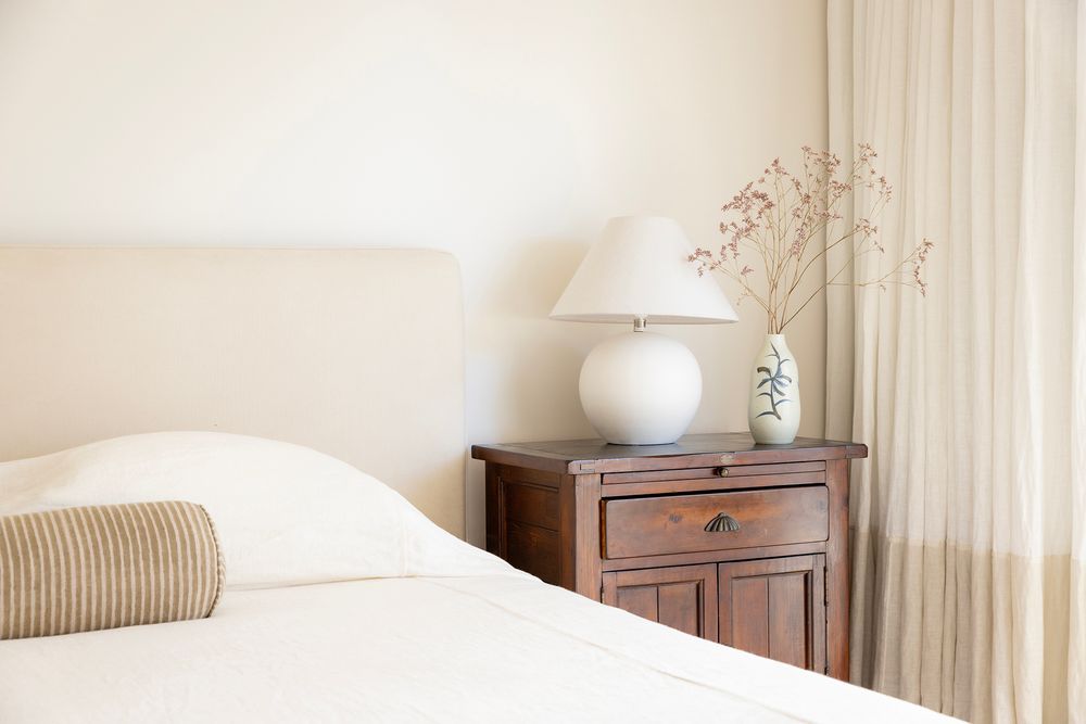 white linen bedspread with lamp and flowers and white curtain in background