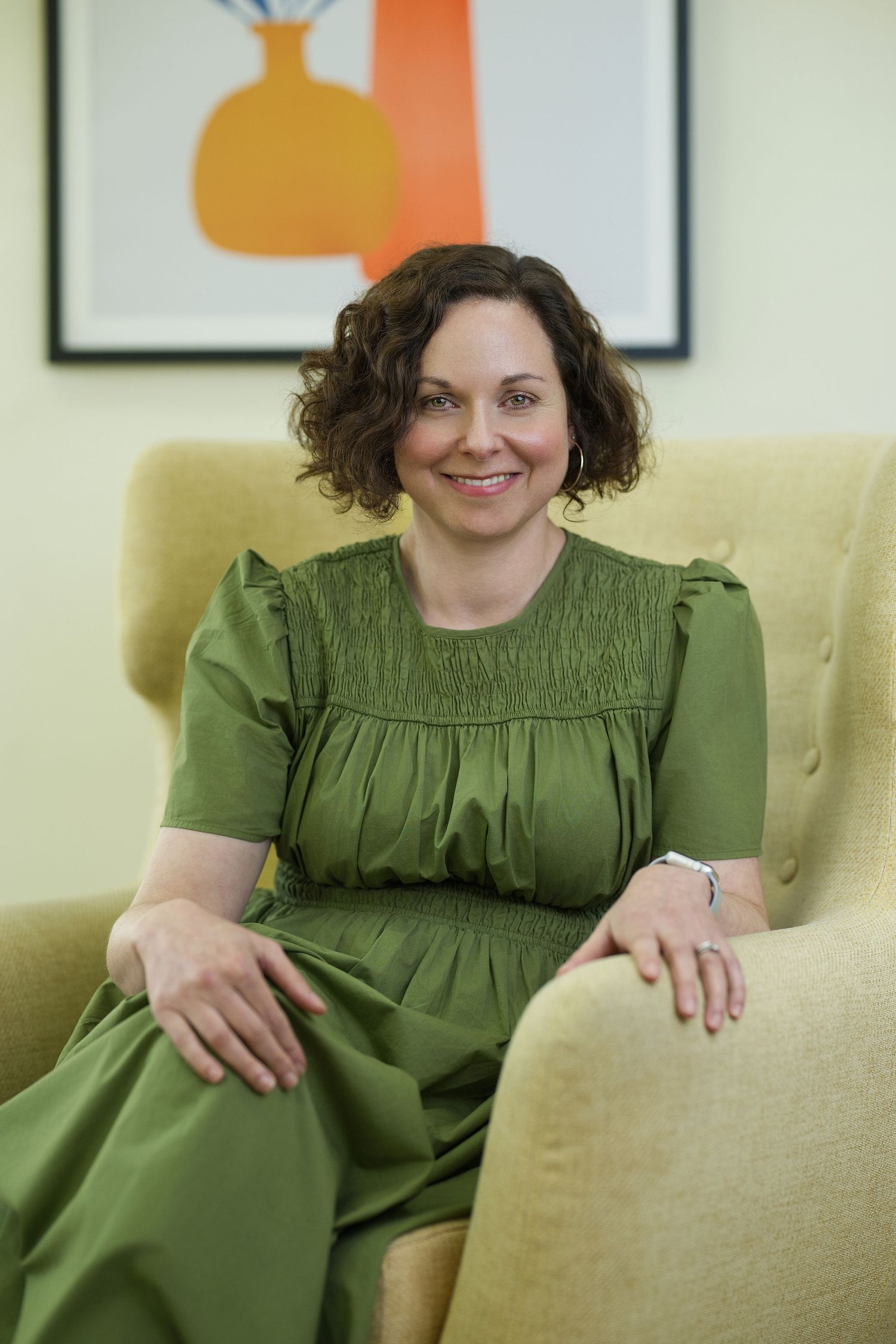Woman wearing a green dress sits comfortably on a cream-coloured chair with modern art on the wall behind. Professional business portrait.