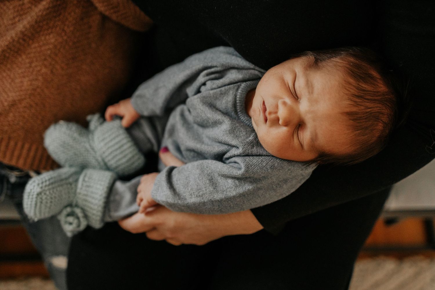 A sleeping baby in gray clothes rests peacefully in someone's arms with warm lighting.