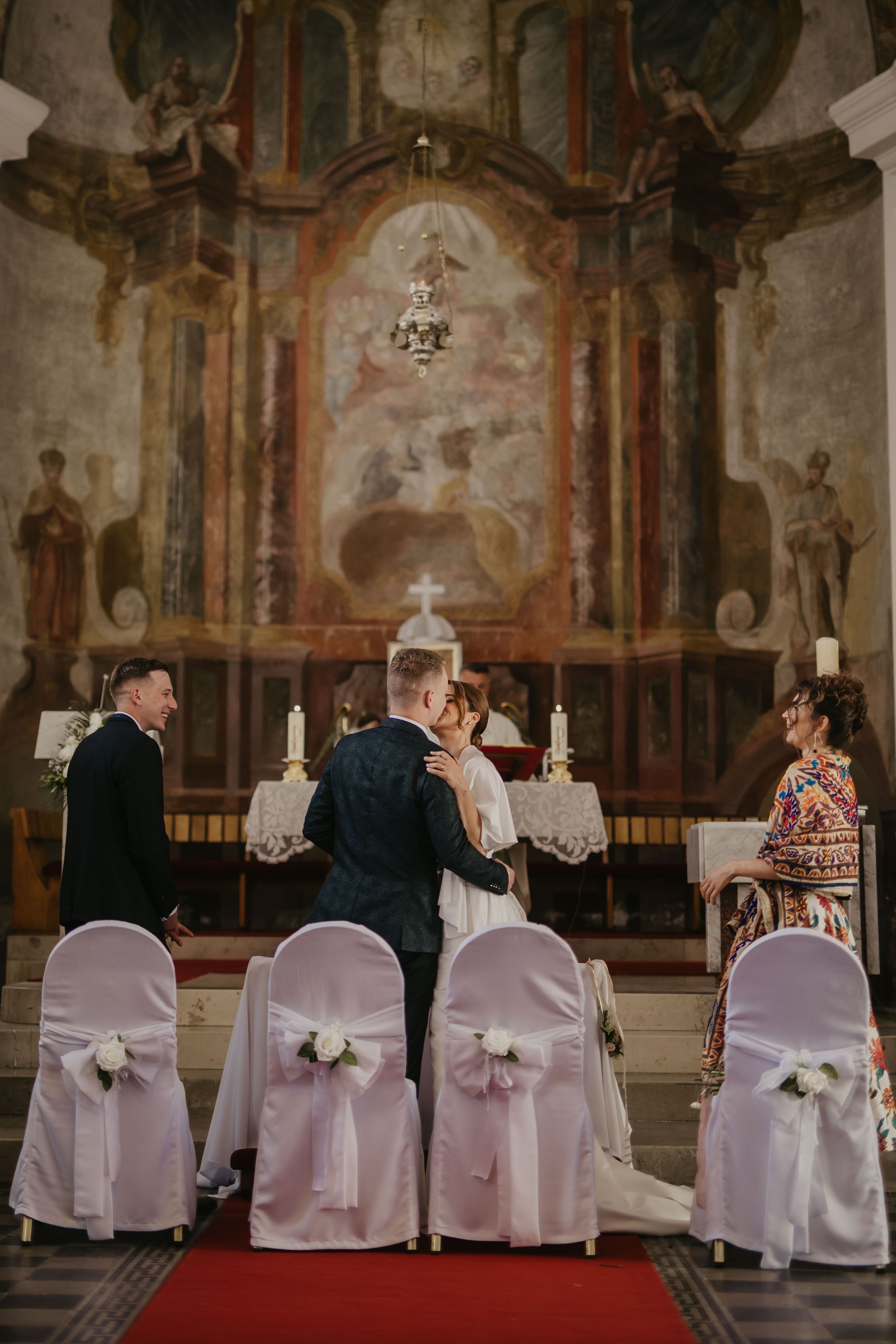 Wedding ceremony in a historic church with baroque altar and white chair covers adorned with bows.