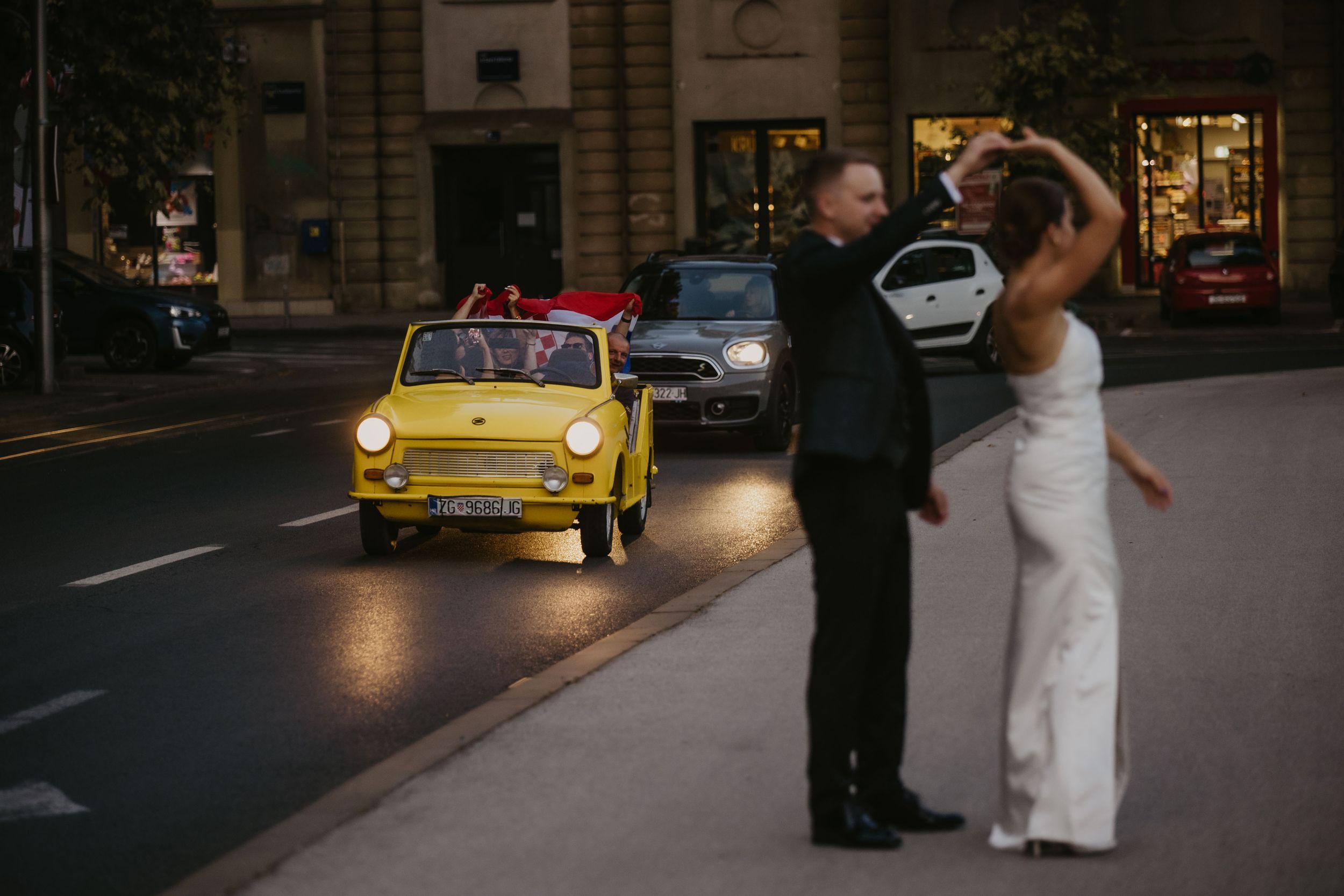Wedding couple dancing in front of a yellow vintage Fiat car on a city street at dusk.