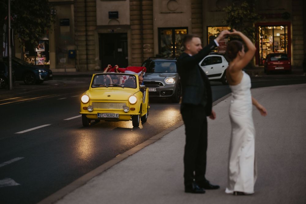 Wedding couple dancing in front of a yellow vintage Fiat car on a city street at dusk.