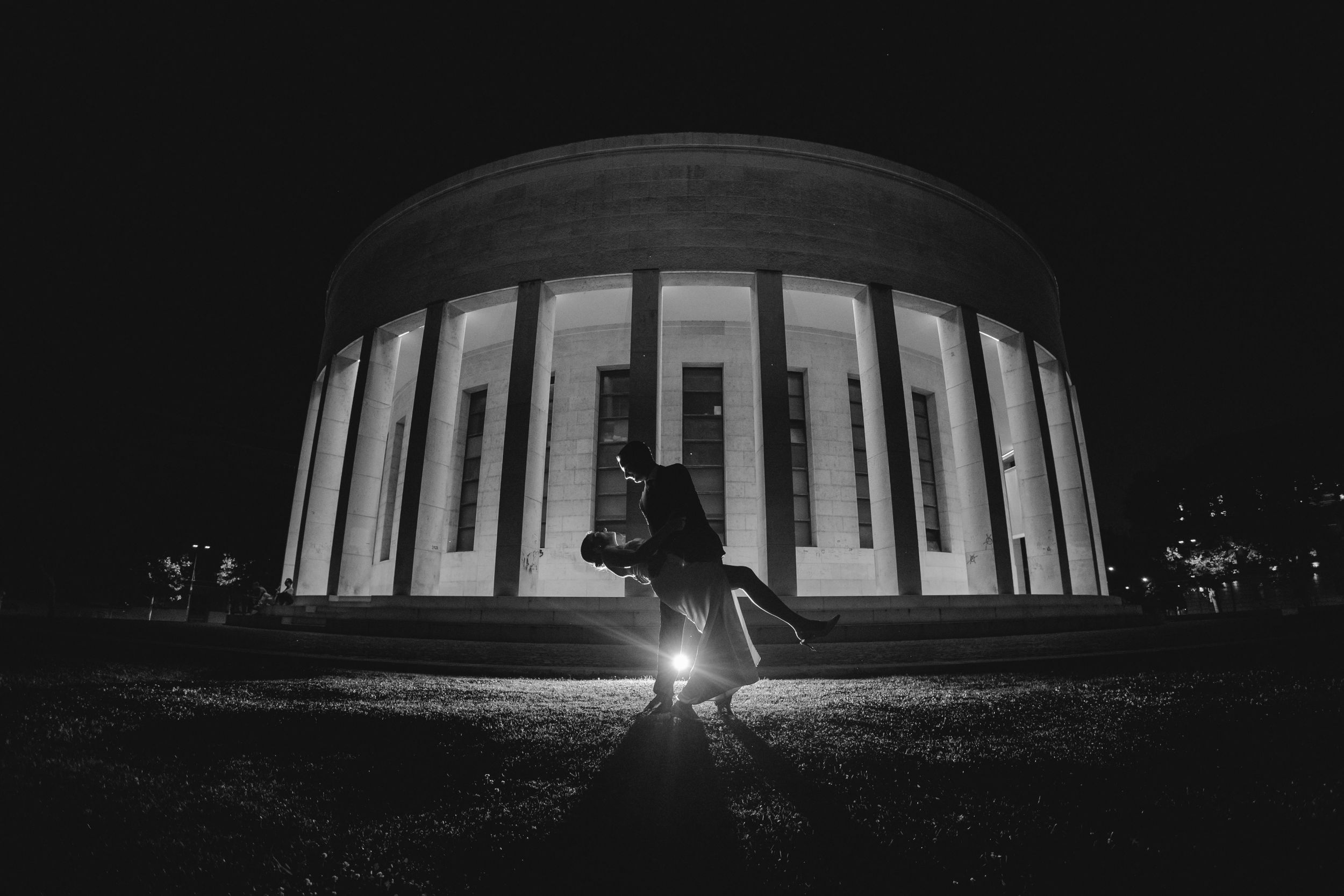 Dramatic nighttime silhouette of a wedding couple in front of an illuminated circular building.