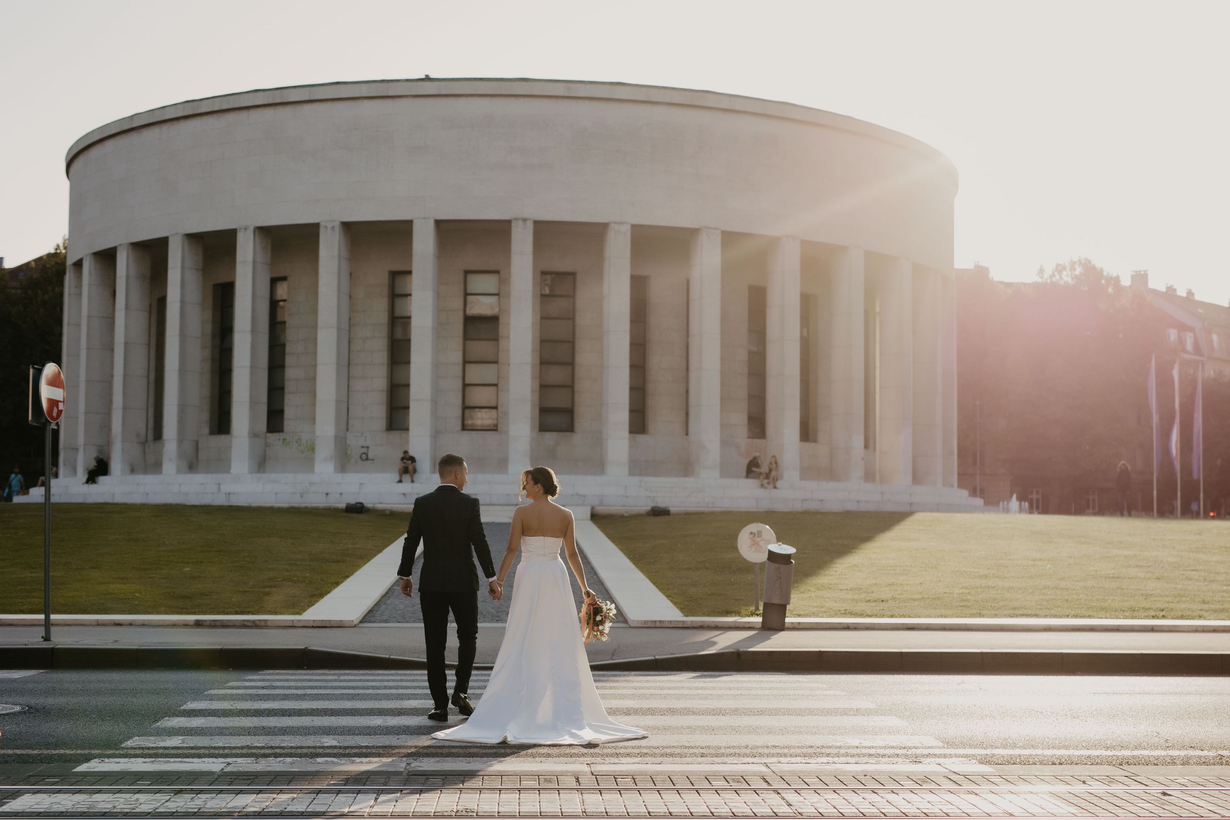 Newlyweds walk hand in hand in front of a grand columned building during sunset.