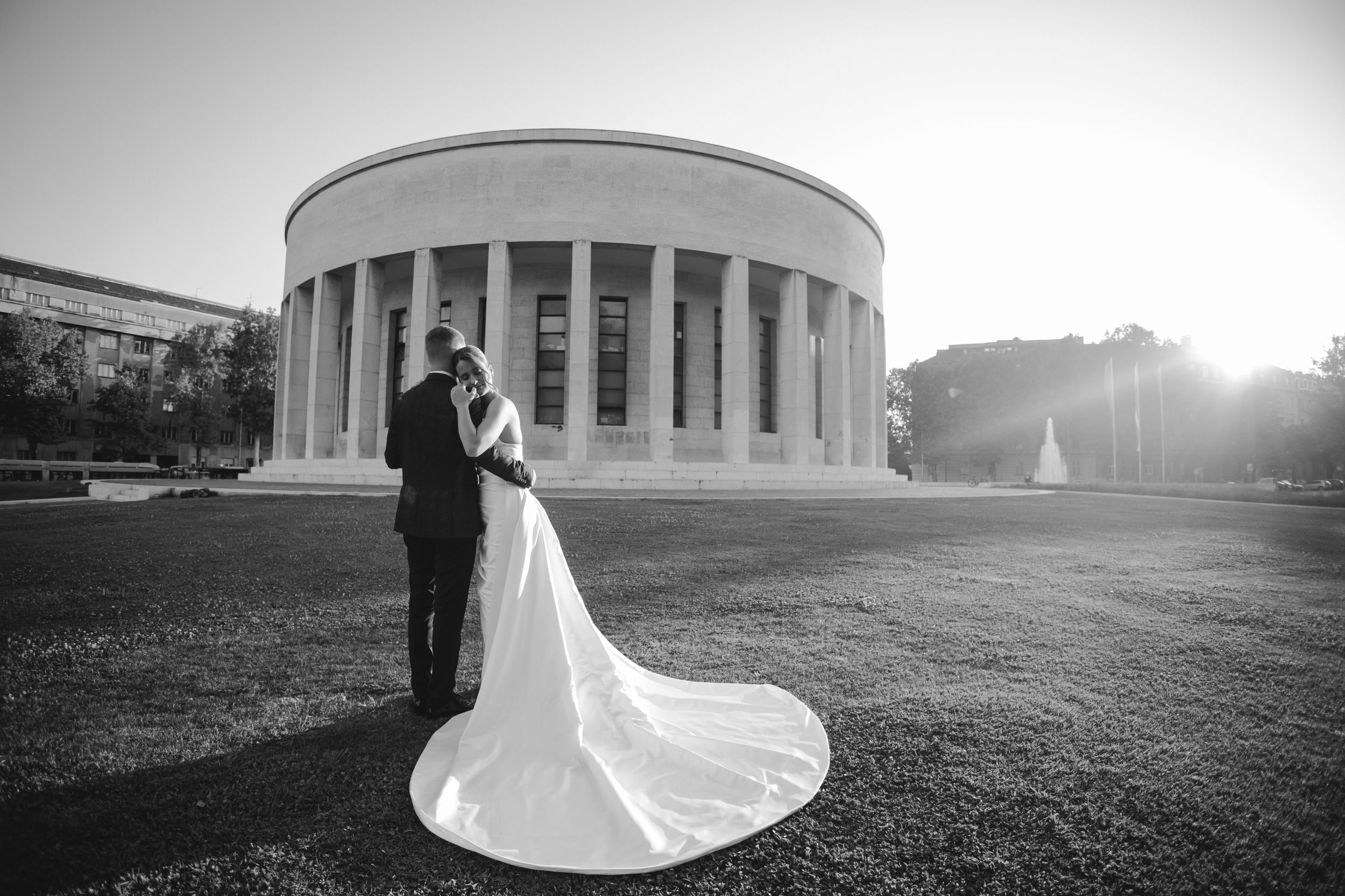 A wedding couple poses in silhouette against a grand columned building at sunset with a flowing white dress train.