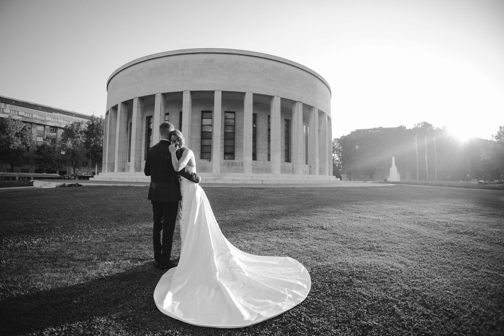 A wedding couple poses in silhouette against a grand columned building at sunset with a flowing white dress train.