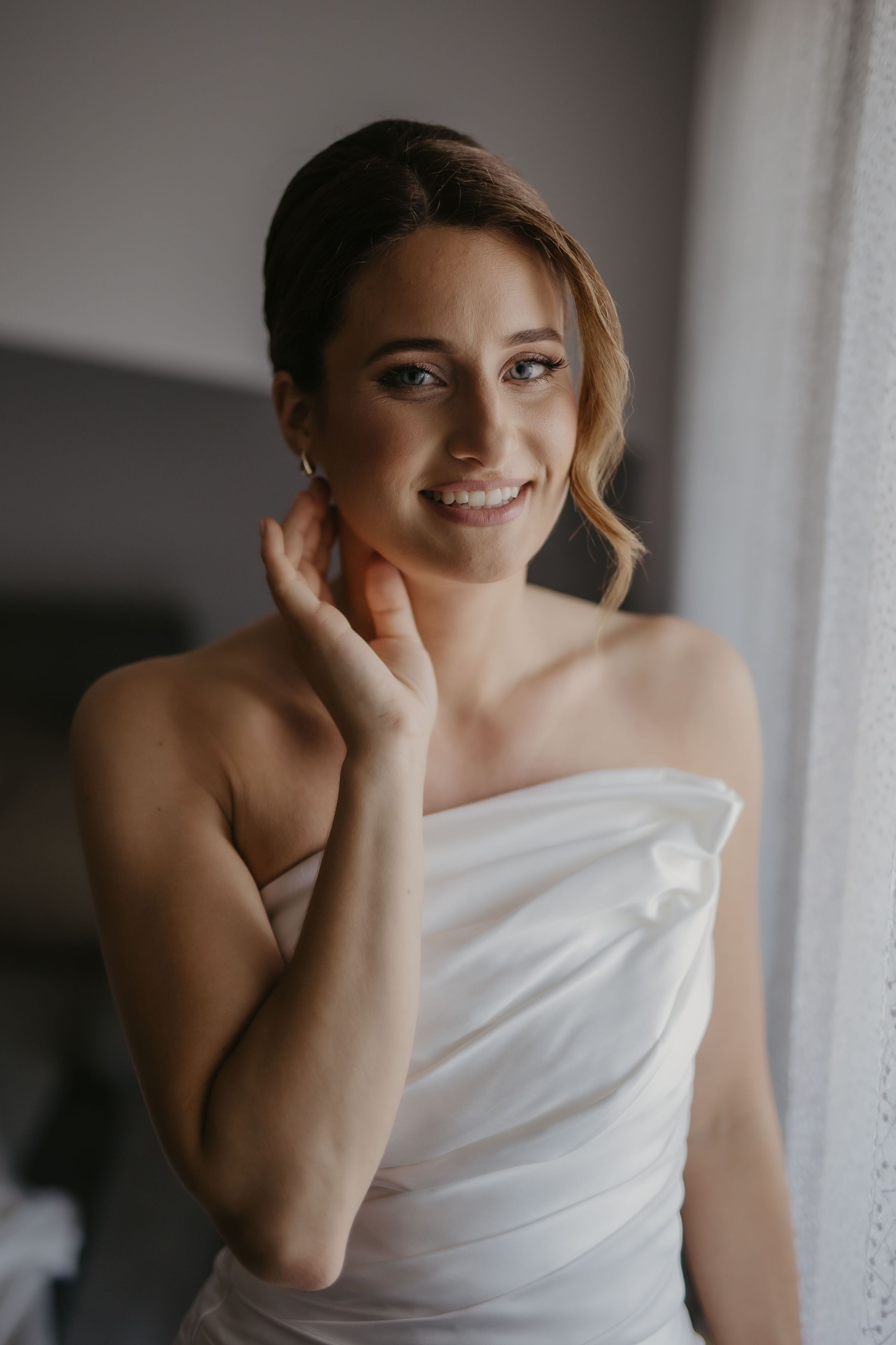 Portrait of someone in a white strapless wedding dress by a window with soft natural light.