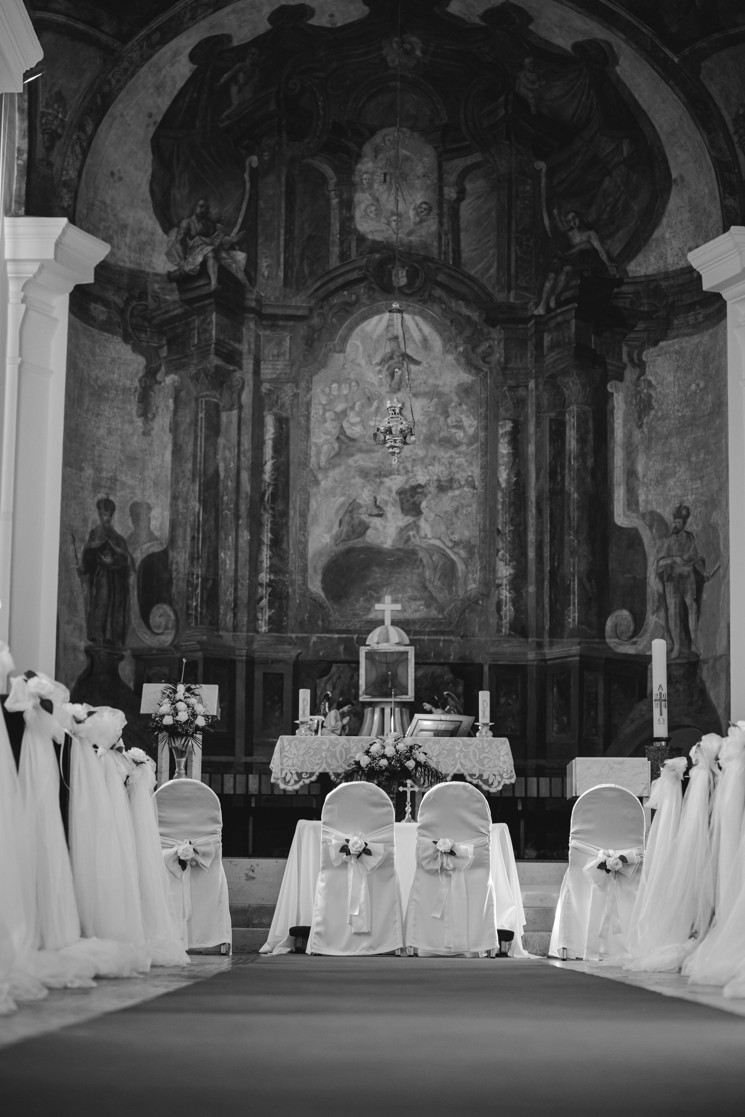 Black and white photograph of a church altar decorated with white chair covers for a wedding ceremony.