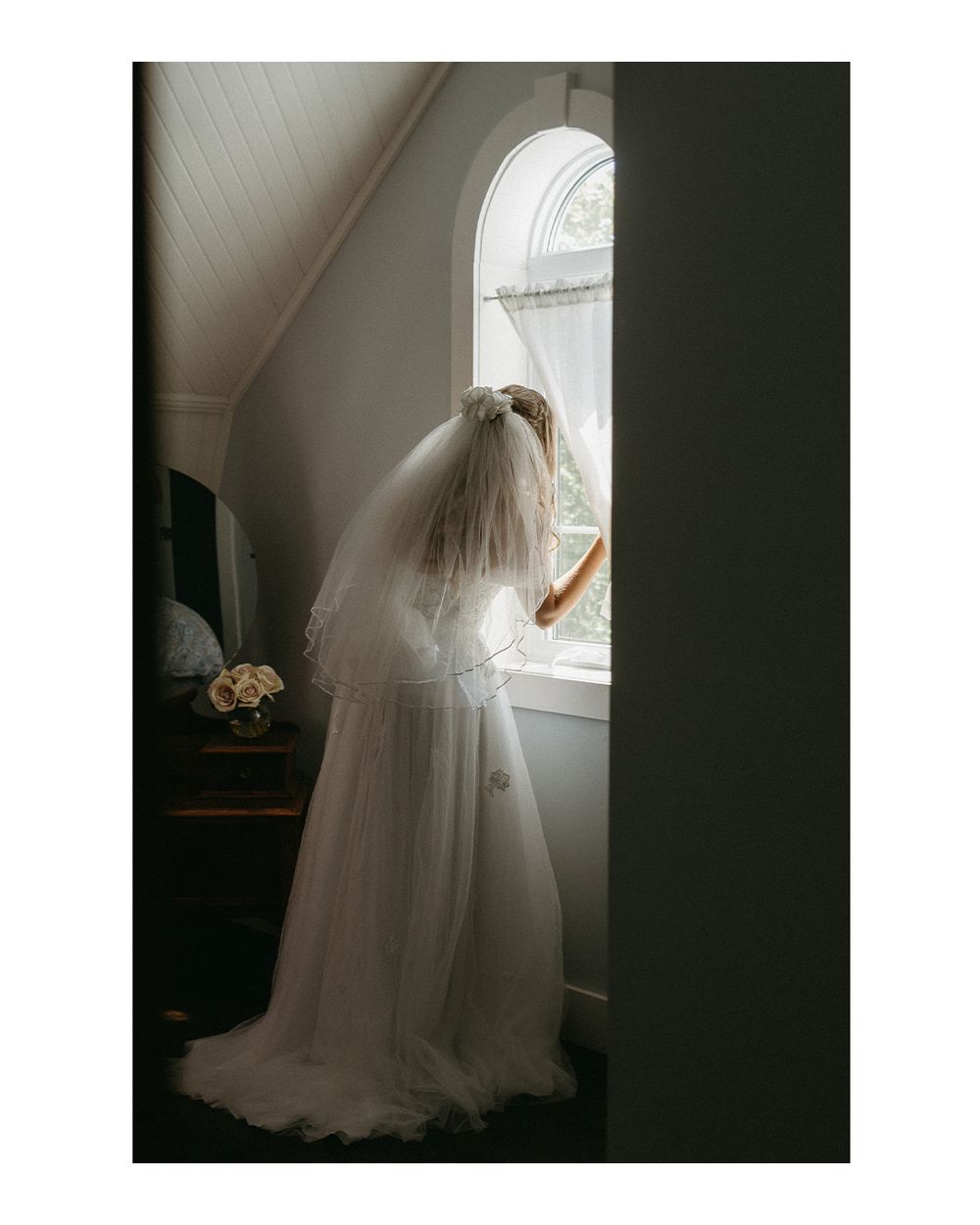 A figure in a flowing white wedding dress and veil stands silhouetted against a bright window in a moody setting.