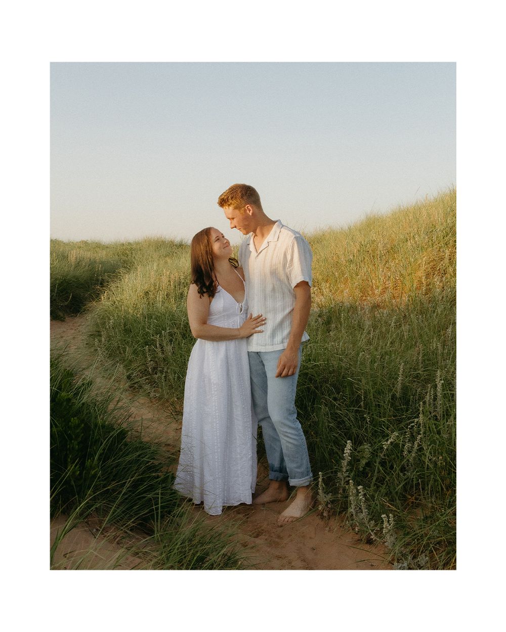 A couple shares a romantic moment in a golden field at sunset while wearing white and denim outfits.