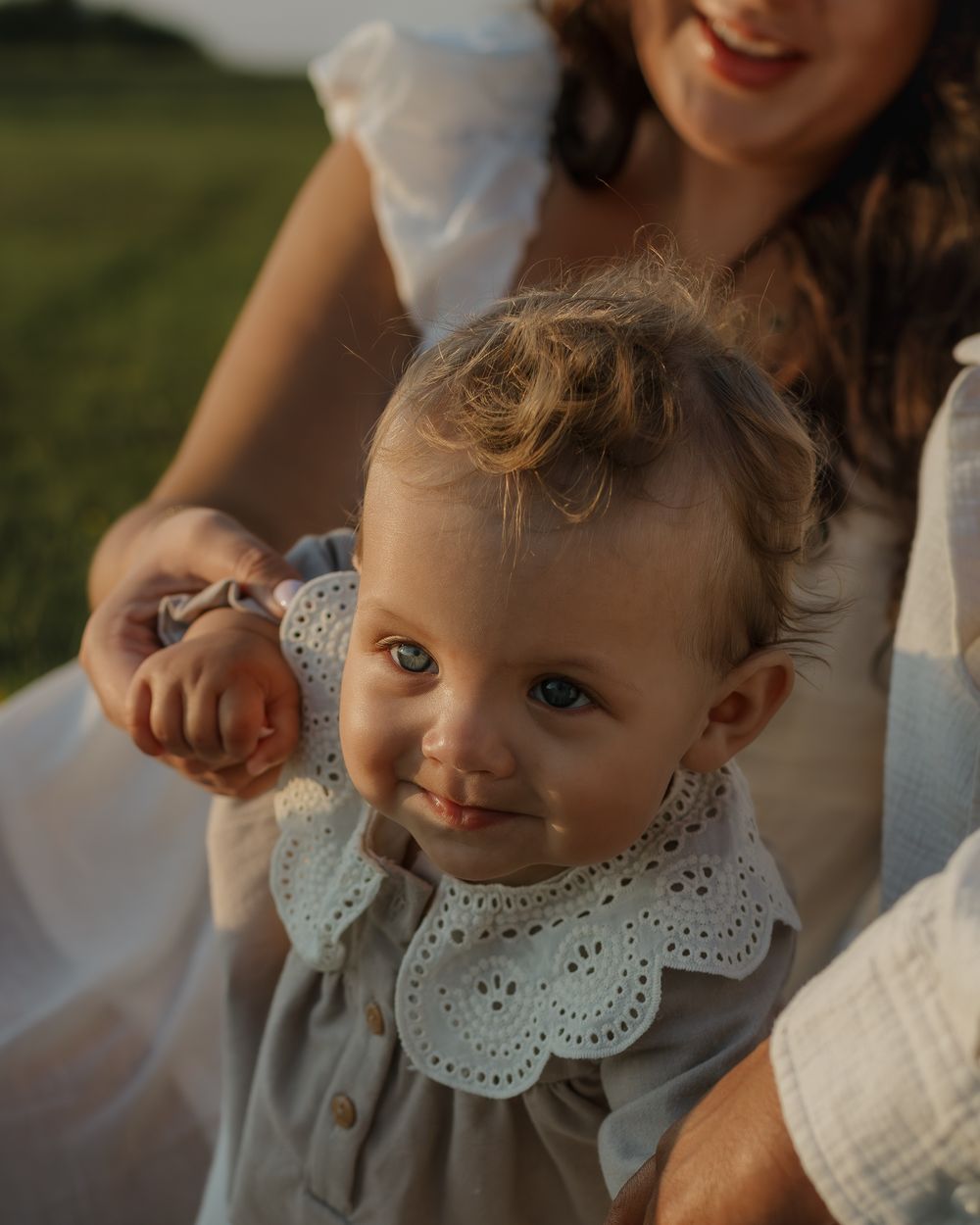 A toddler wearing a gray outfit with a white collar smiles brightly in outdoor golden light.