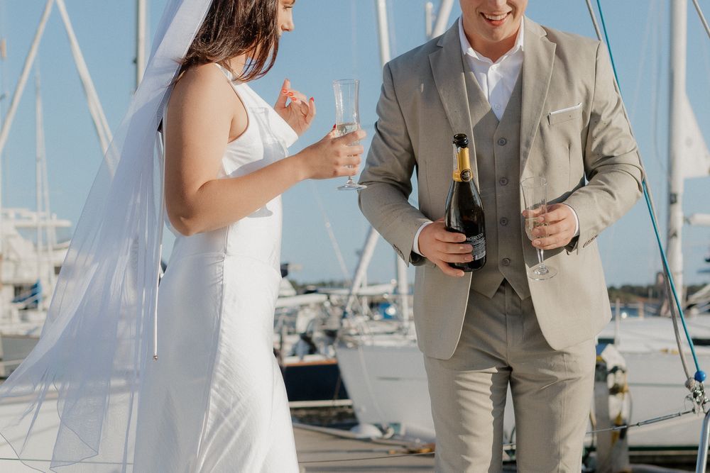 A couple in wedding attire share drinks on a dock with sailboats in the marina background.