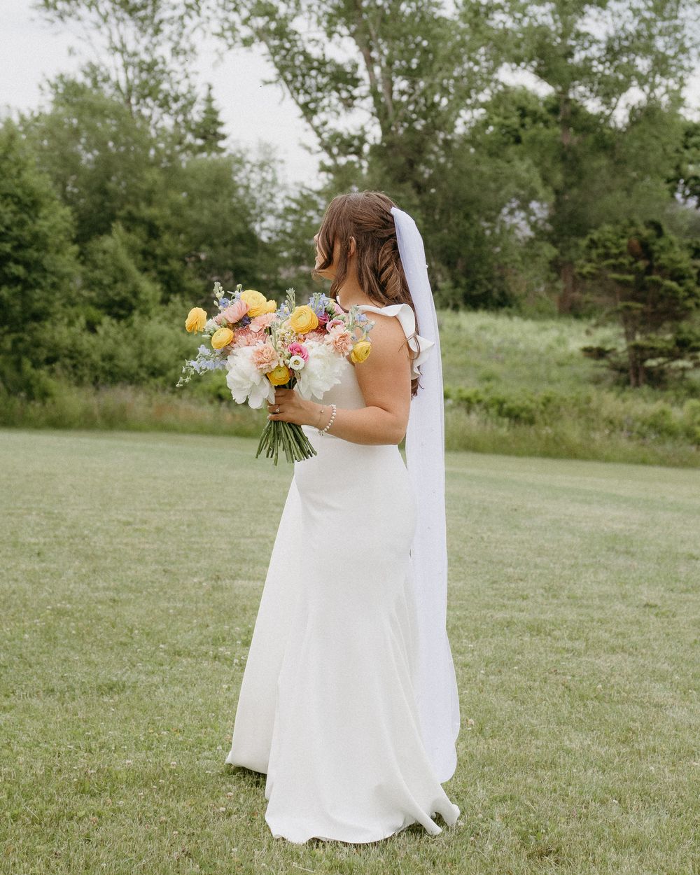 A bride in a flowing white dress holds a pastel bouquet while standing in a lush green meadow on a sunny day.