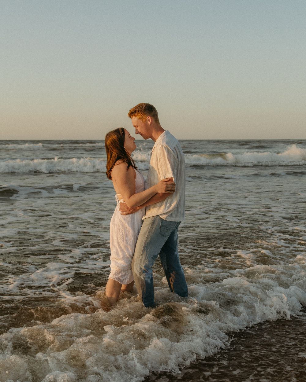 A couple shares a romantic moment while standing in the ocean waves at sunset, their silhouettes framed by the golden light.